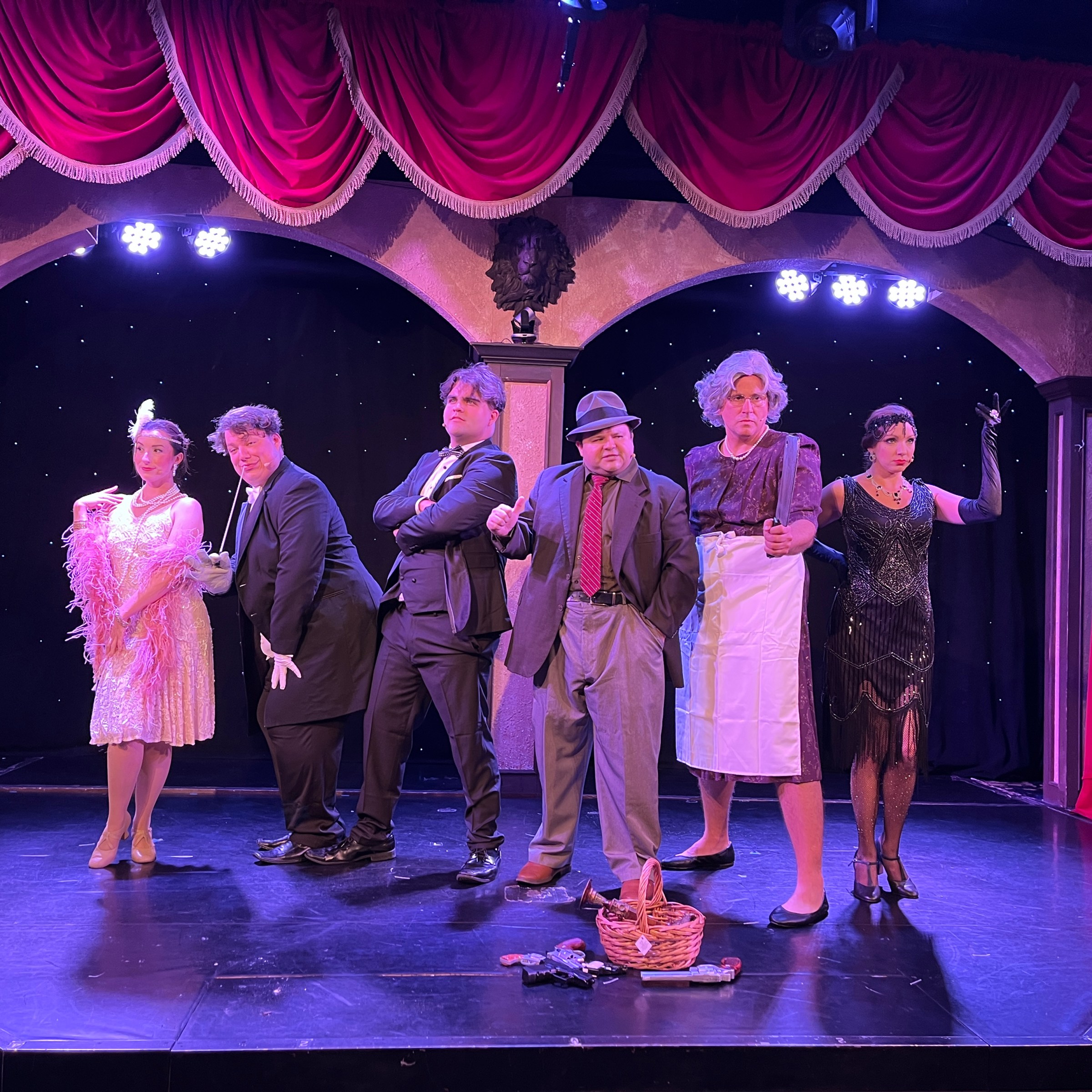 Six people in vintage costumes on stage, red curtains in background.