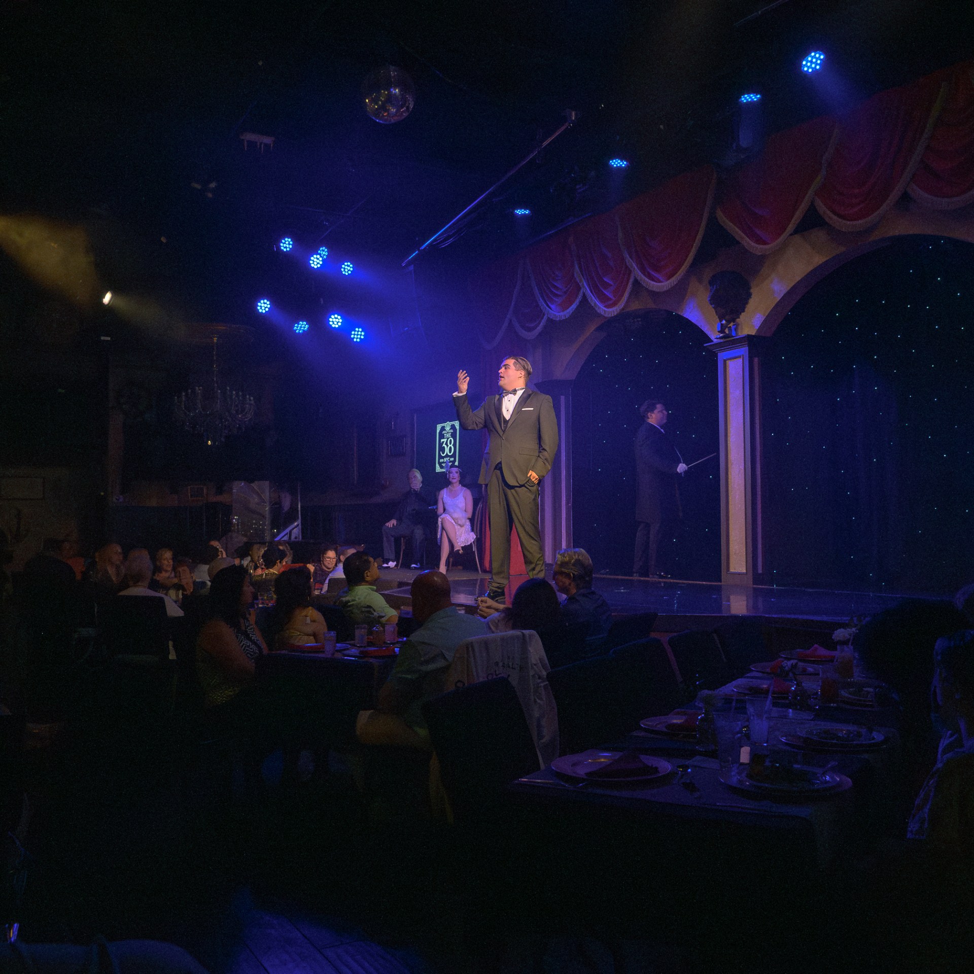 Performers on stage in a dimly lit dinner theater with an audience seated at tables.