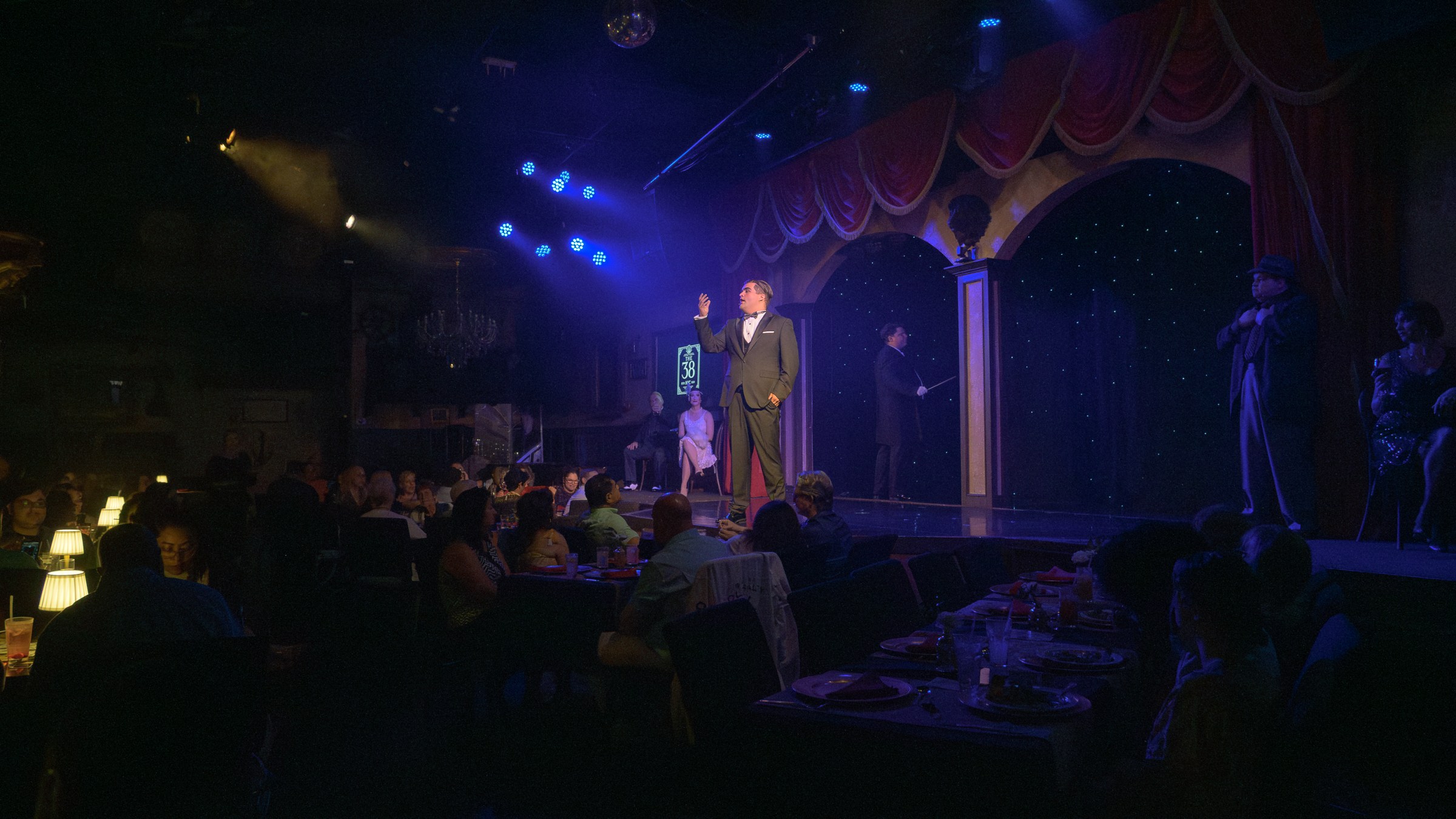 Performers on stage in a dimly lit dinner theater with an audience seated at tables.