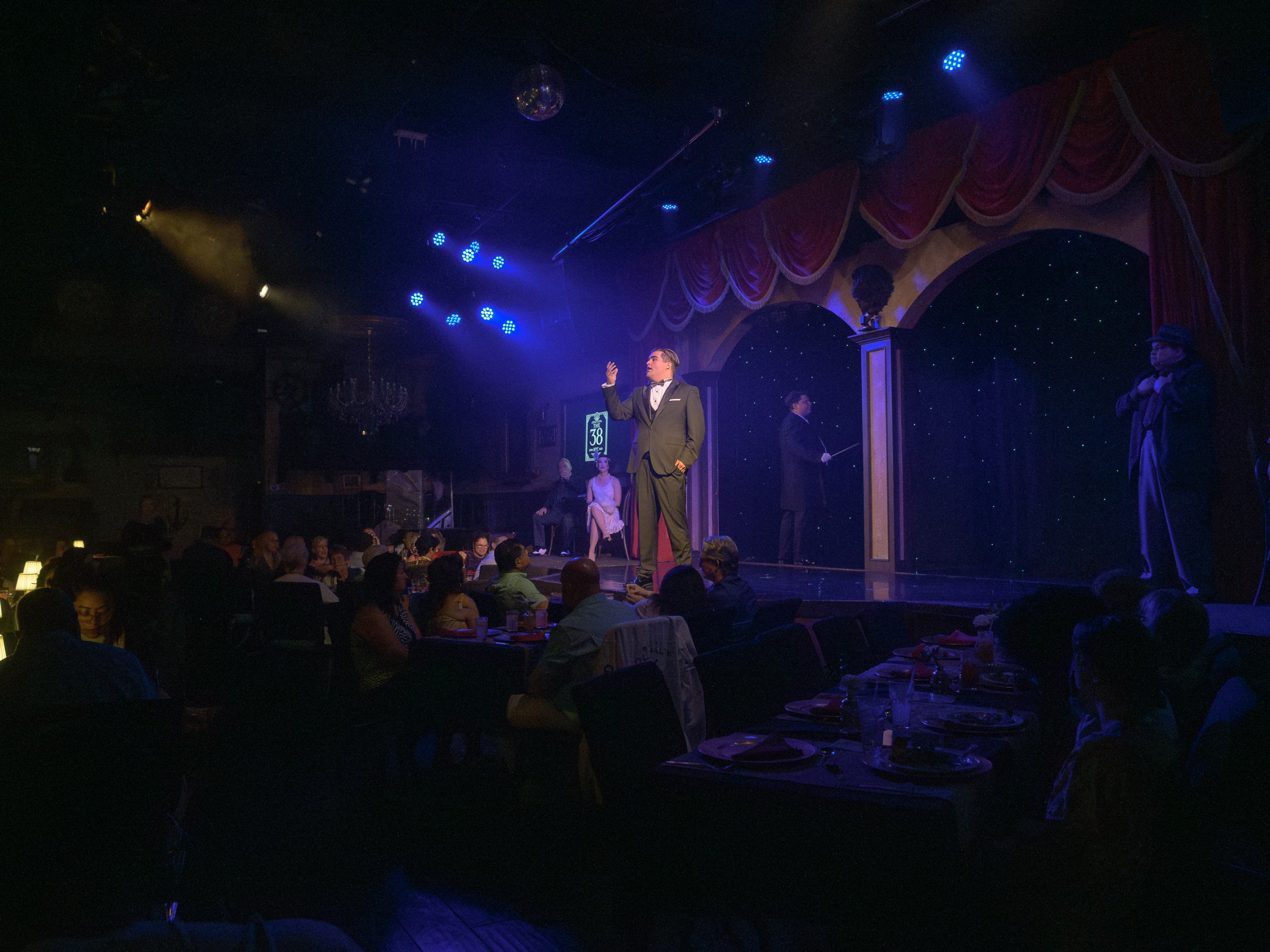 Performers on stage in a dimly lit dinner theater with an audience seated at tables.