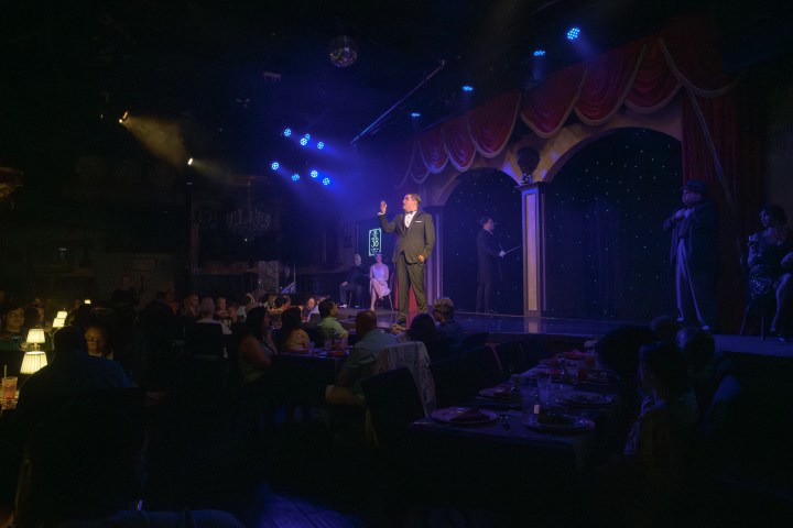 Performers on stage in a dimly lit dinner theater with an audience seated at tables.