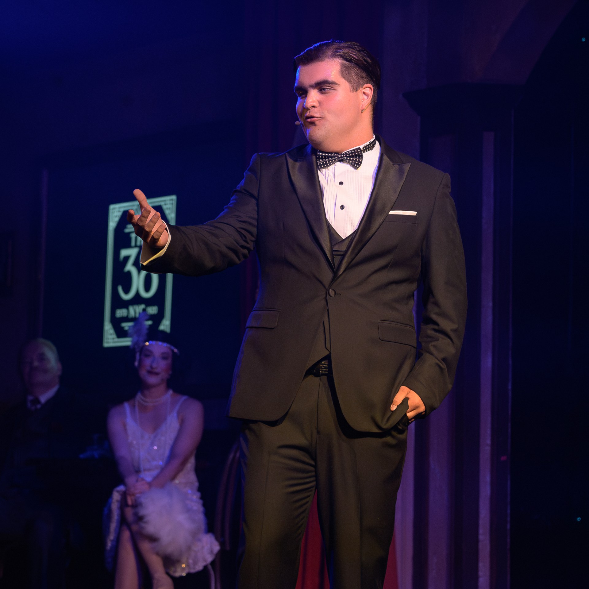 Man in black tuxedo performing on stage with seated audience in dim lighting.