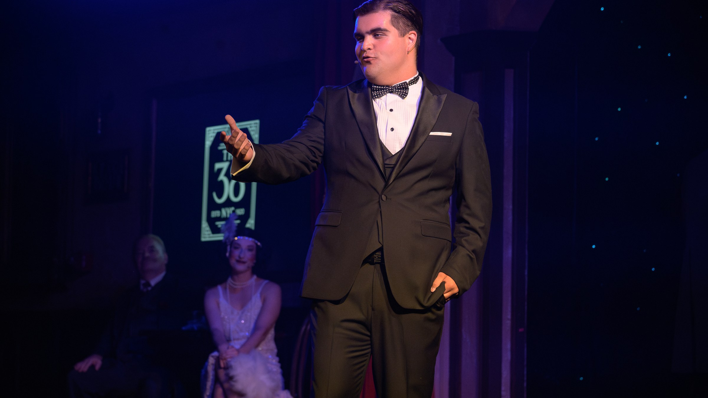 Man in black tuxedo performing on stage with seated audience in dim lighting.