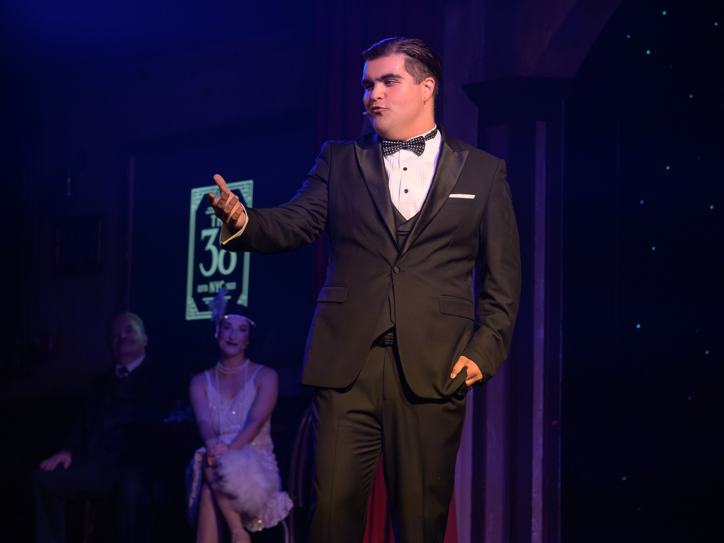 Man in black tuxedo performing on stage with seated audience in dim lighting.