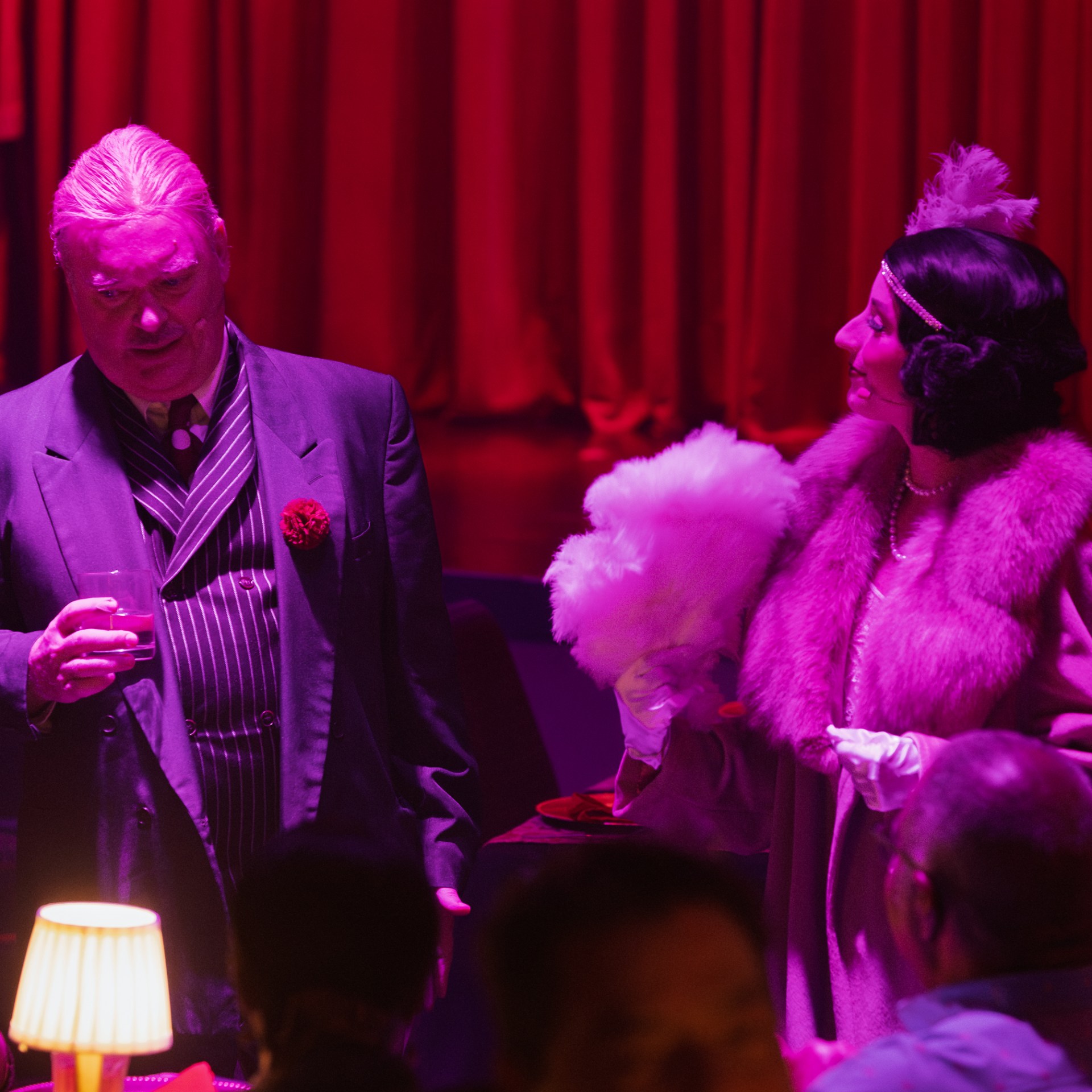 Elegantly dressed man and woman in vintage style at a dimly-lit, red-themed event.