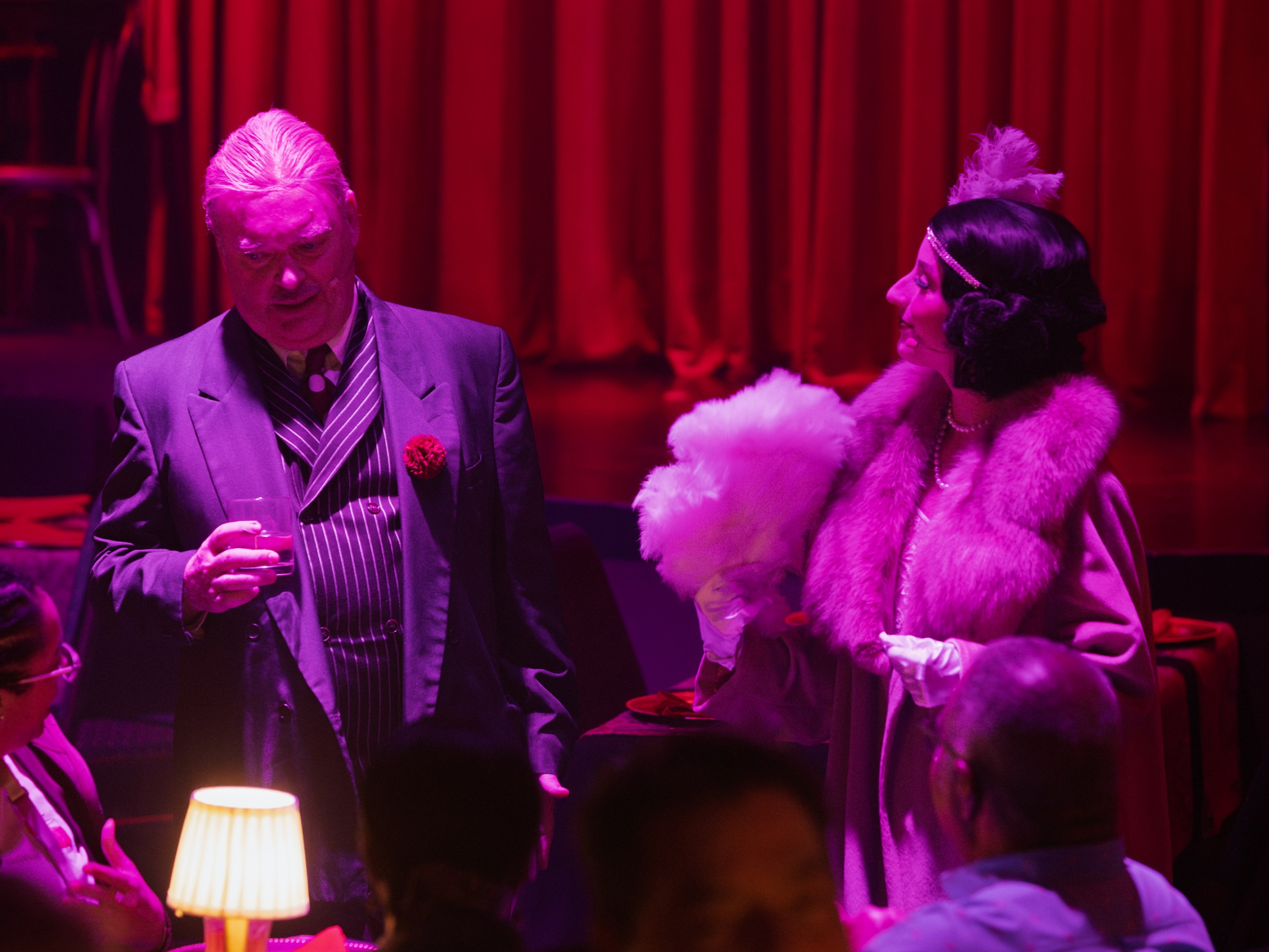 Elegantly dressed man and woman in vintage style at a dimly-lit, red-themed event.