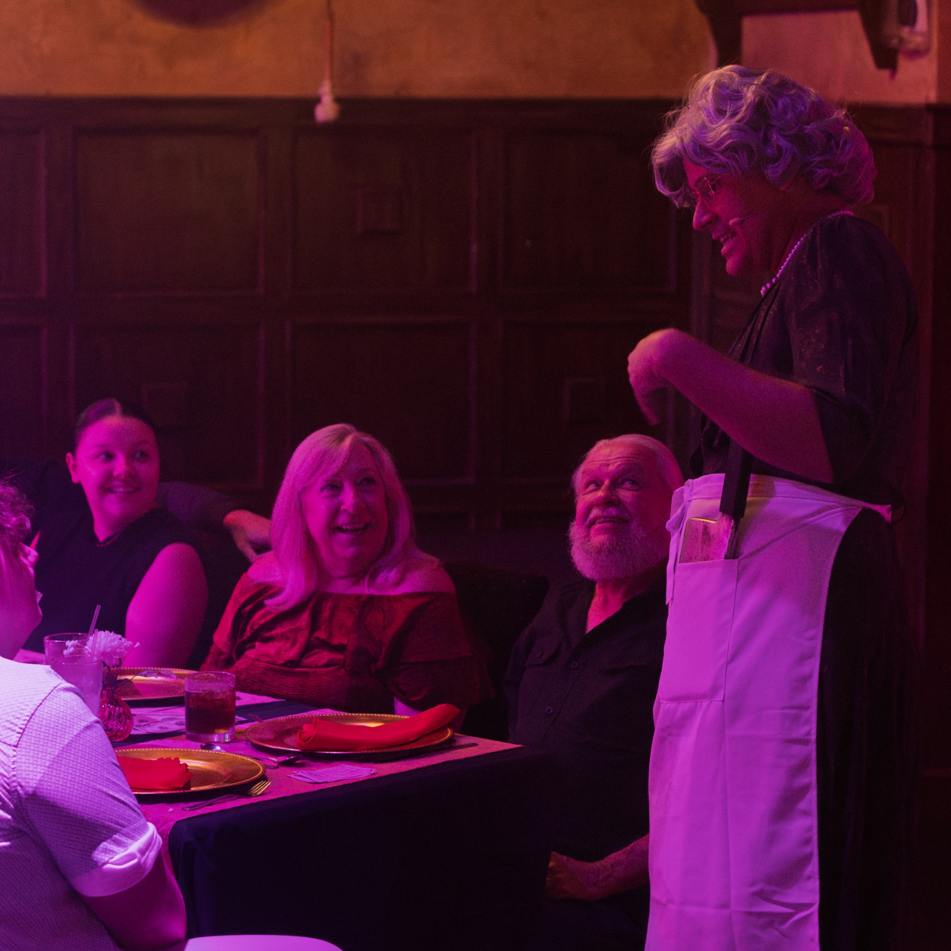 Person in costume entertaining a group seated at a dining table with colorful lighting.