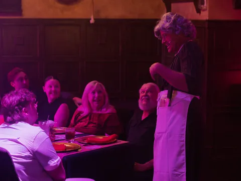 Person in costume entertaining a group seated at a dining table with colorful lighting.