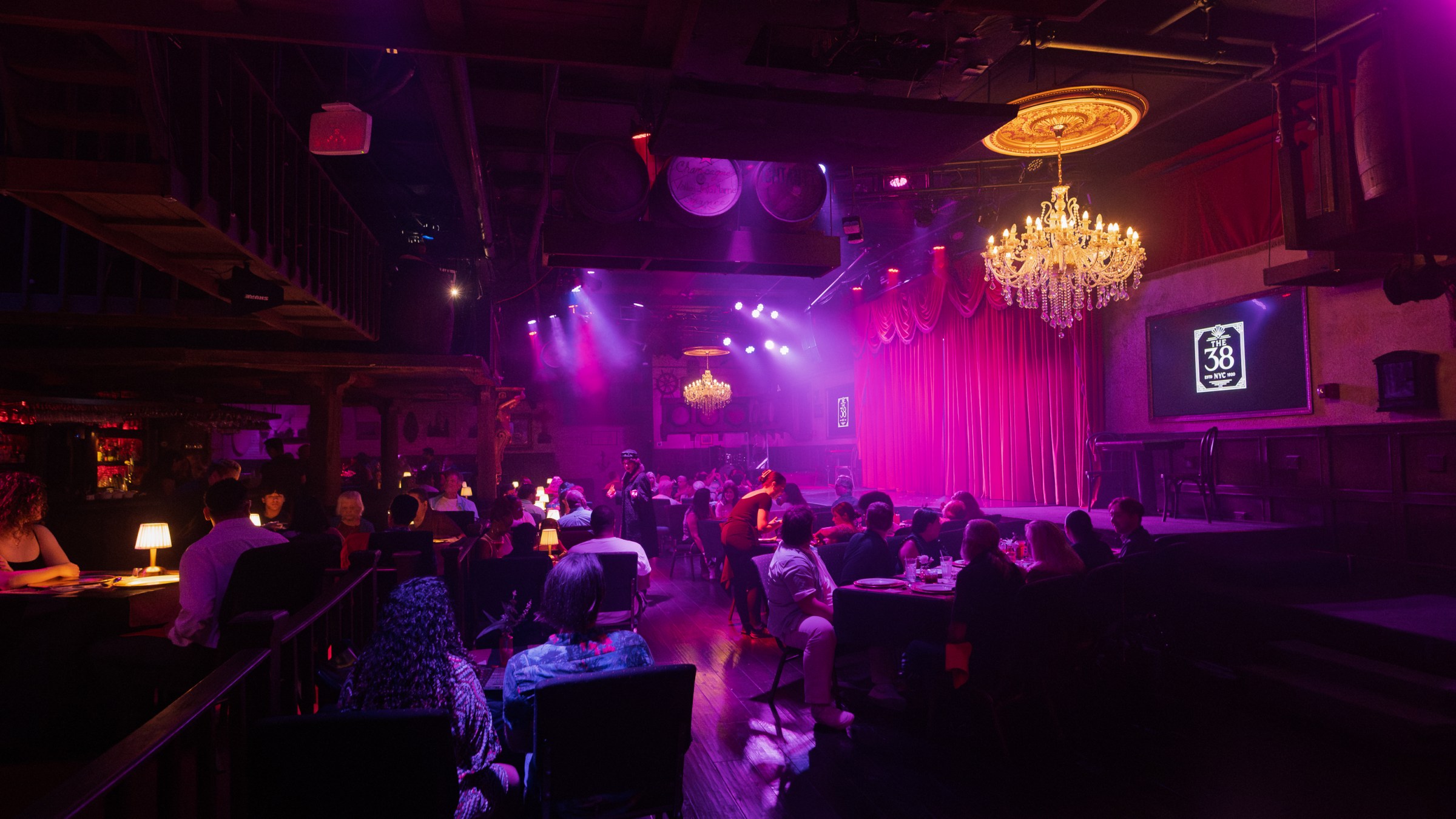 Dimly lit club with chandeliers, an audience seated at tables, and a stage with red curtains.