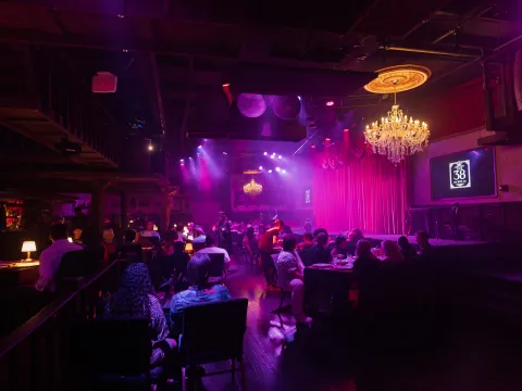 Dimly lit club with chandeliers, an audience seated at tables, and a stage with red curtains.