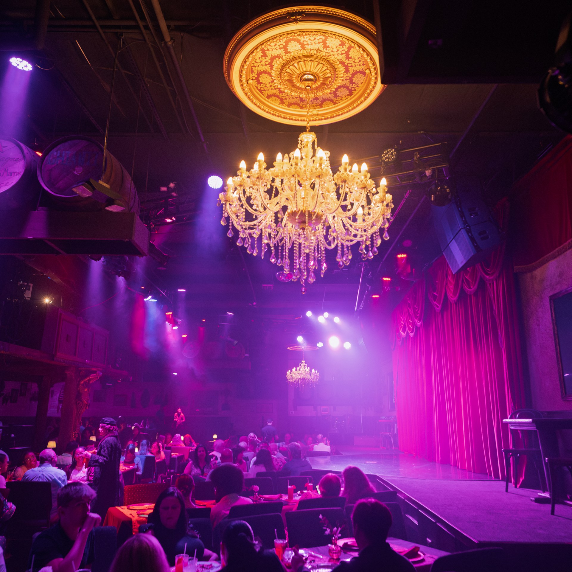 Dimly lit theater with chandeliers, red curtains, and seated audience.