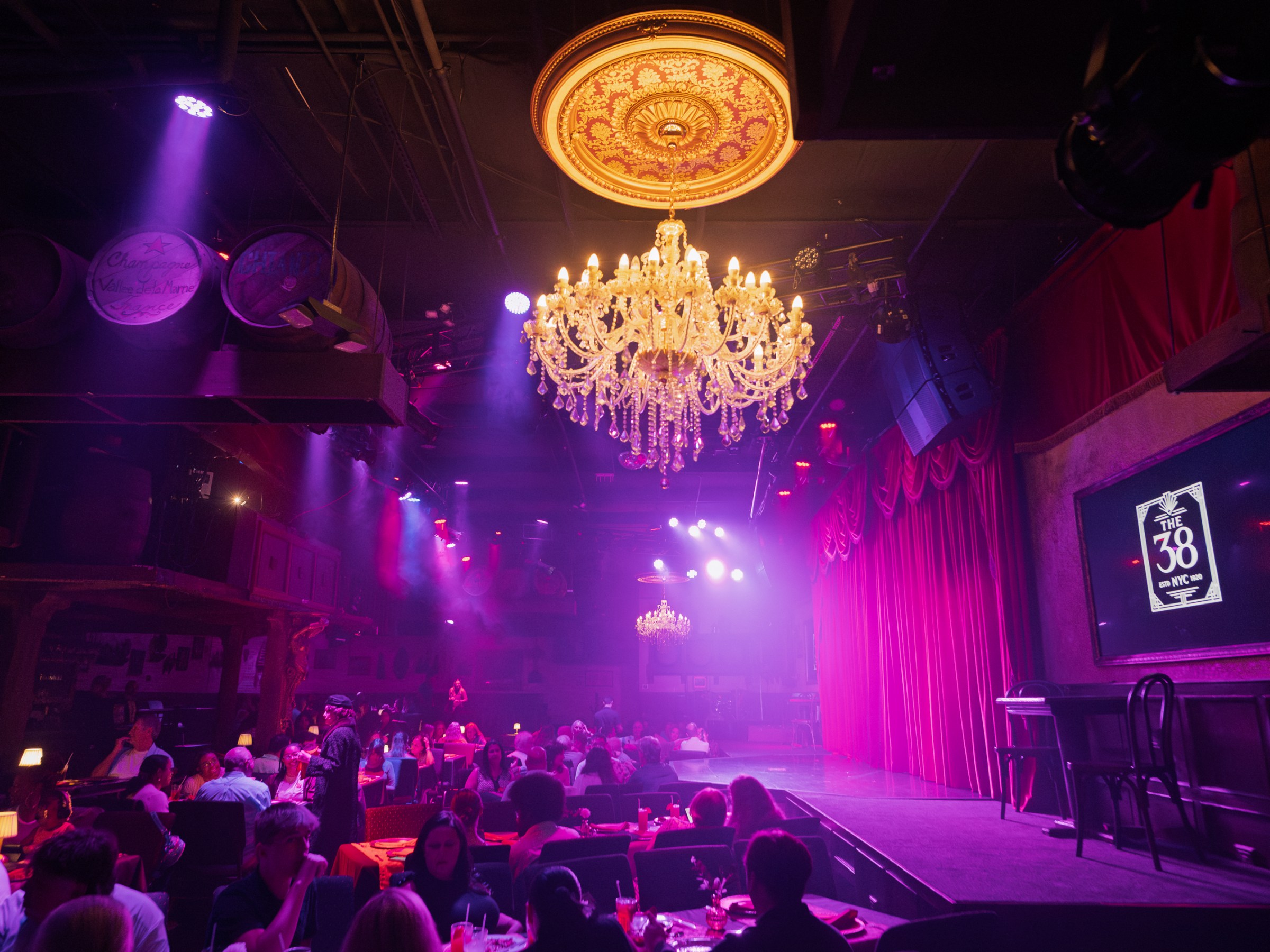 Dimly lit theater with chandeliers, red curtains, and seated audience.