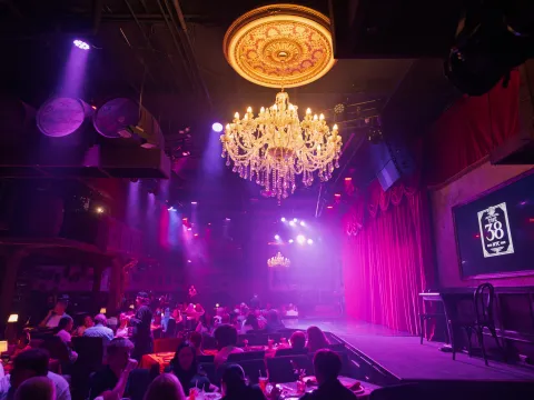 Dimly lit theater with chandeliers, red curtains, and seated audience.
