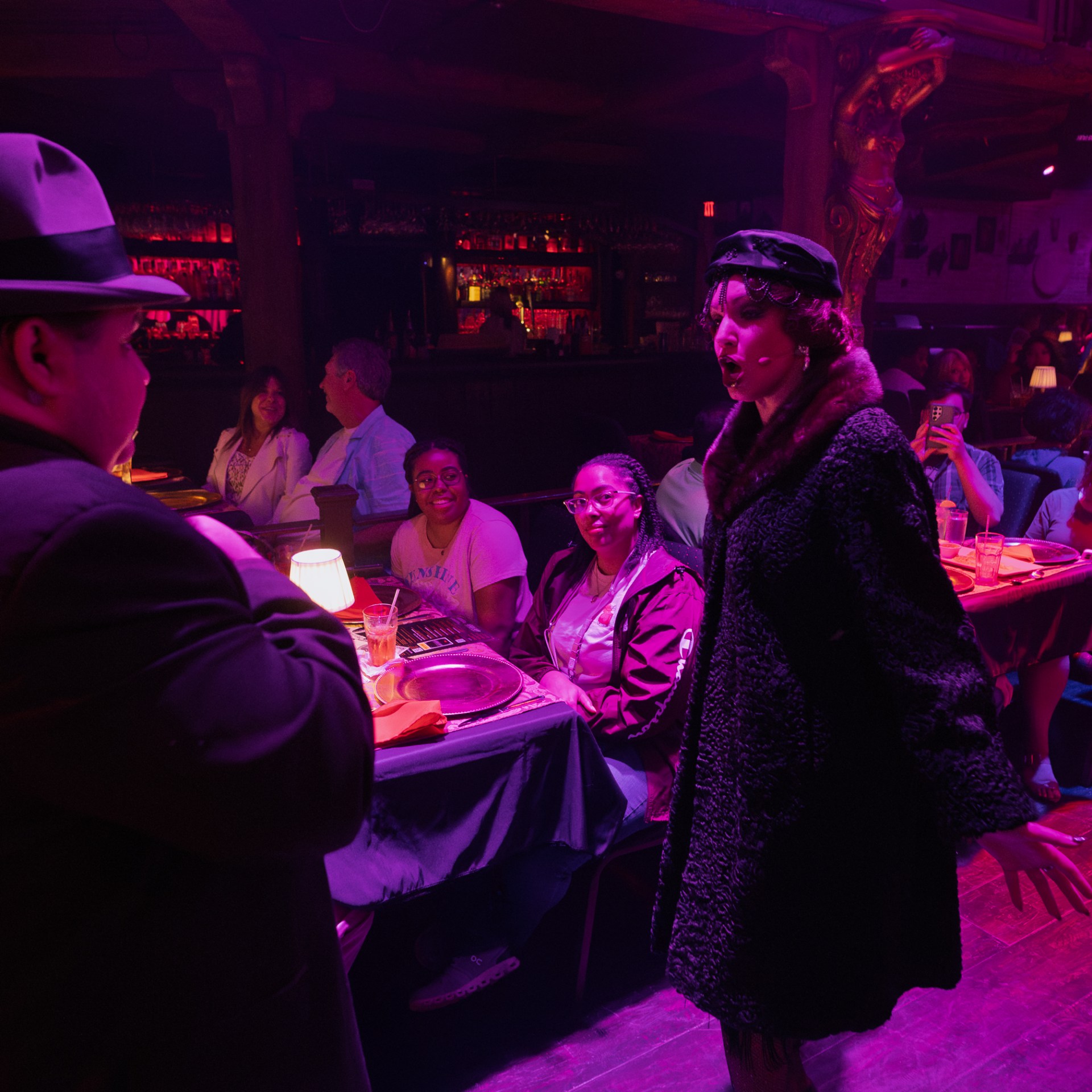 Performers in vintage attire interact with a seated audience in a dimly lit, atmospheric venue.