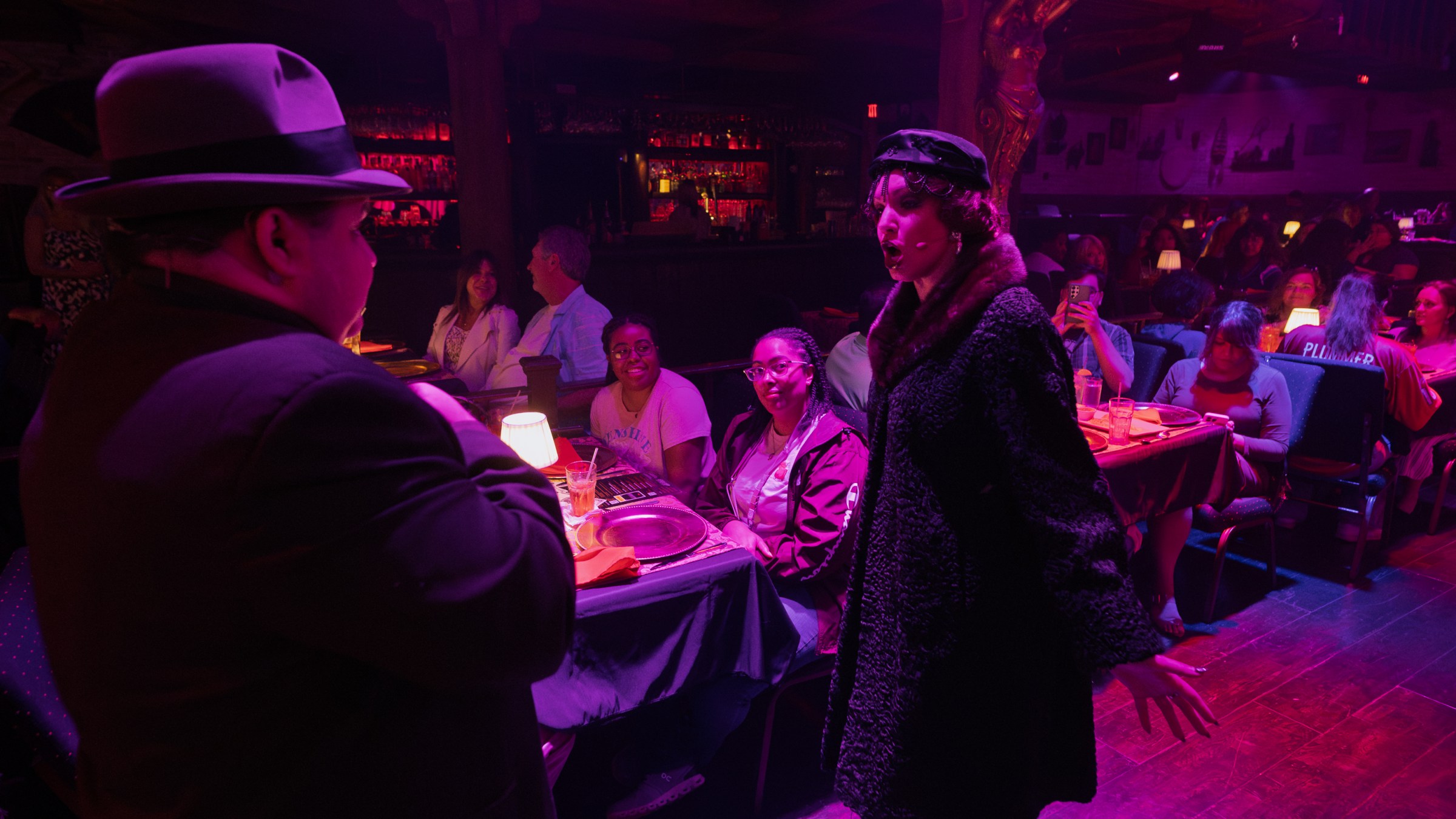 Performers in vintage attire interact with a seated audience in a dimly lit, atmospheric venue.