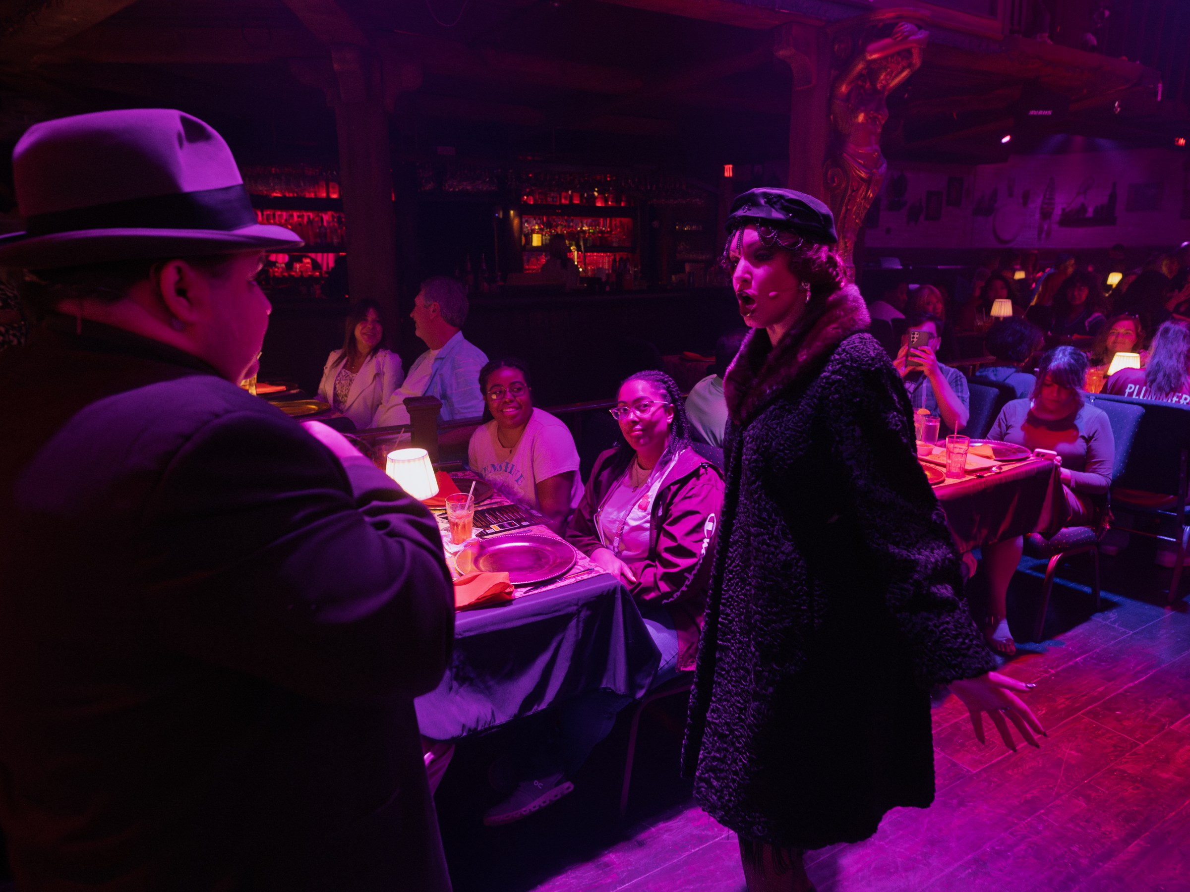 Performers in vintage attire interact with a seated audience in a dimly lit, atmospheric venue.