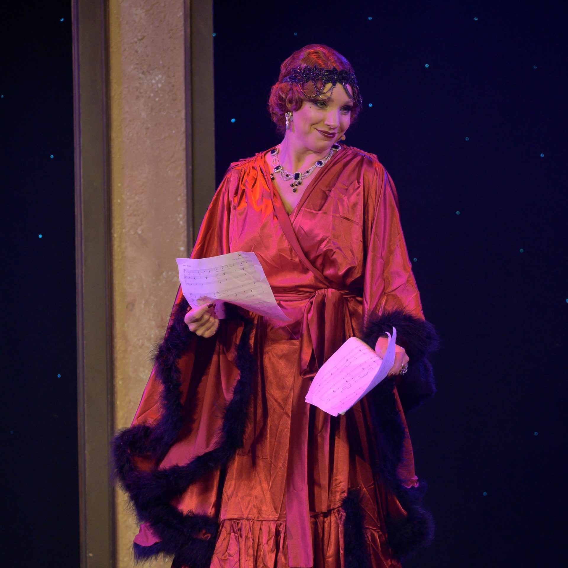 Person in red, vintage-style dress holding sheet music on stage, under stage lights.