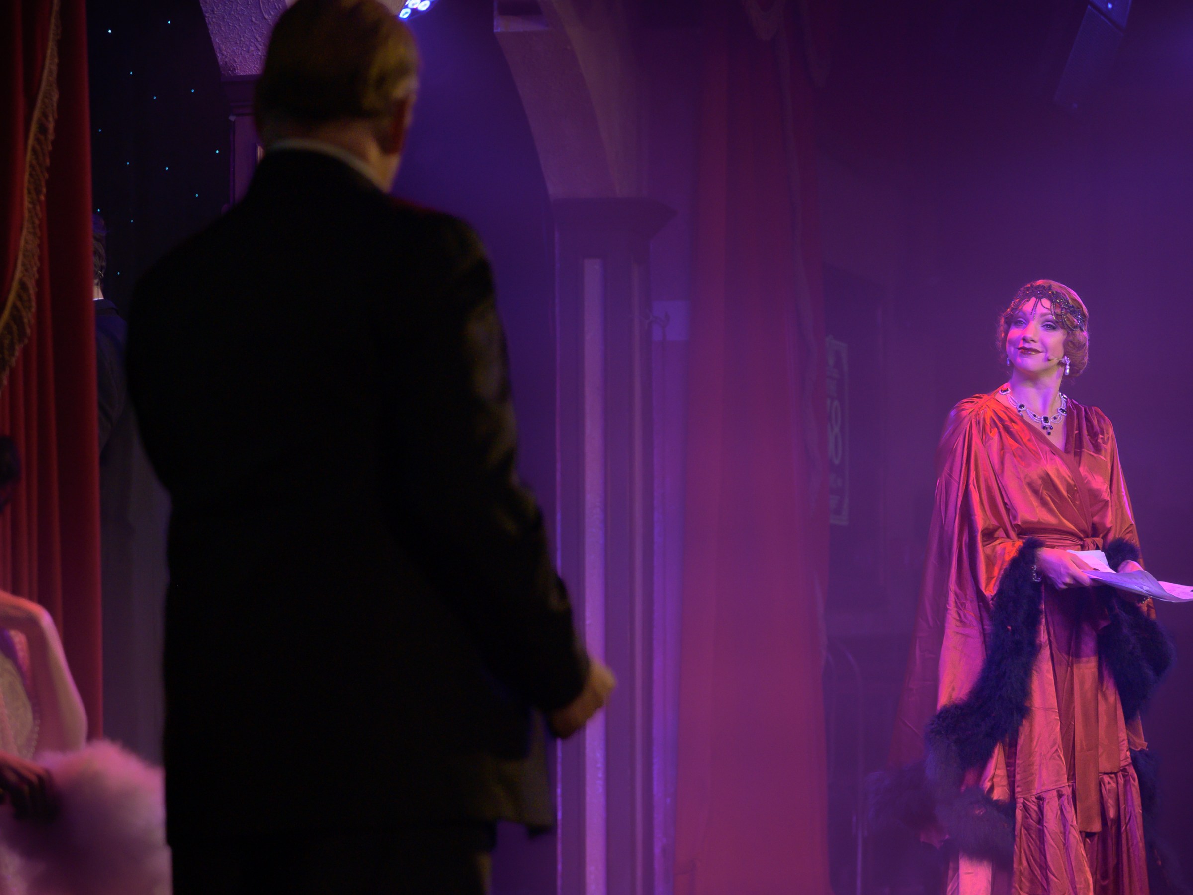 Woman in 1920s attire on stage with man in suit, dimly lit theater.