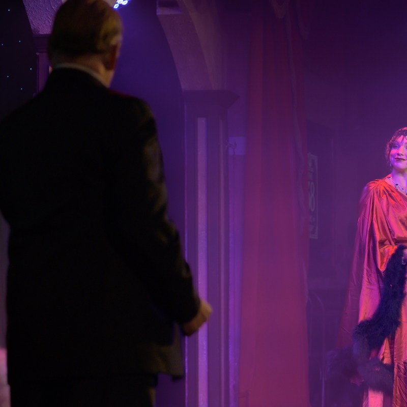 Woman in 1920s attire on stage with man in suit, dimly lit theater.