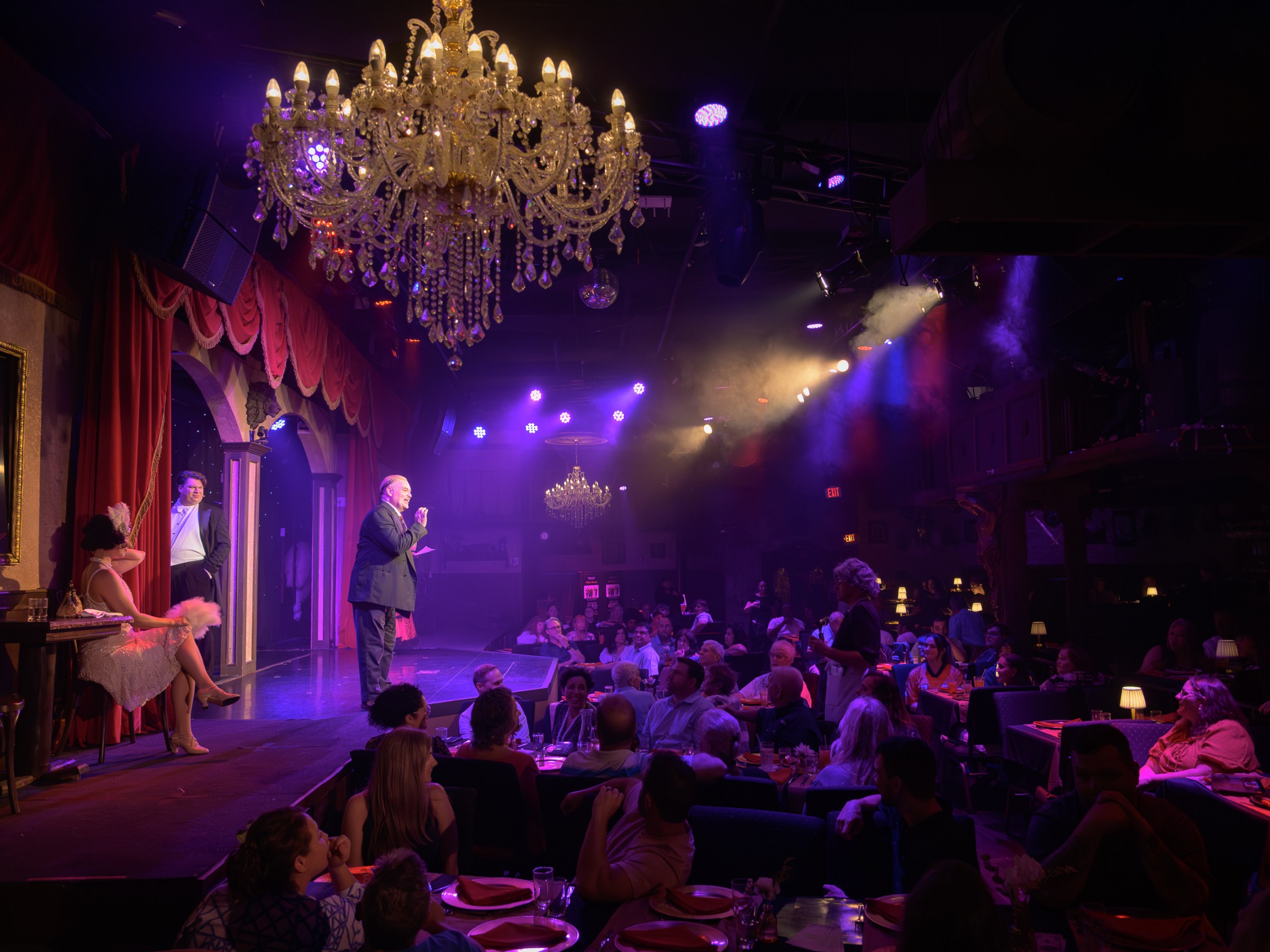 Dimly lit stage performance with audience, chandeliers, and theatrical lighting.