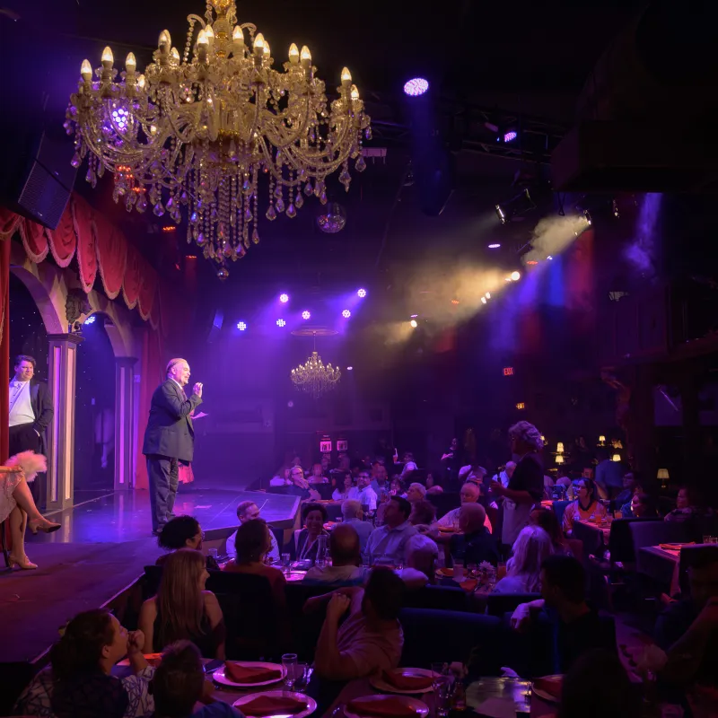 Dimly lit stage performance with audience, chandeliers, and theatrical lighting.