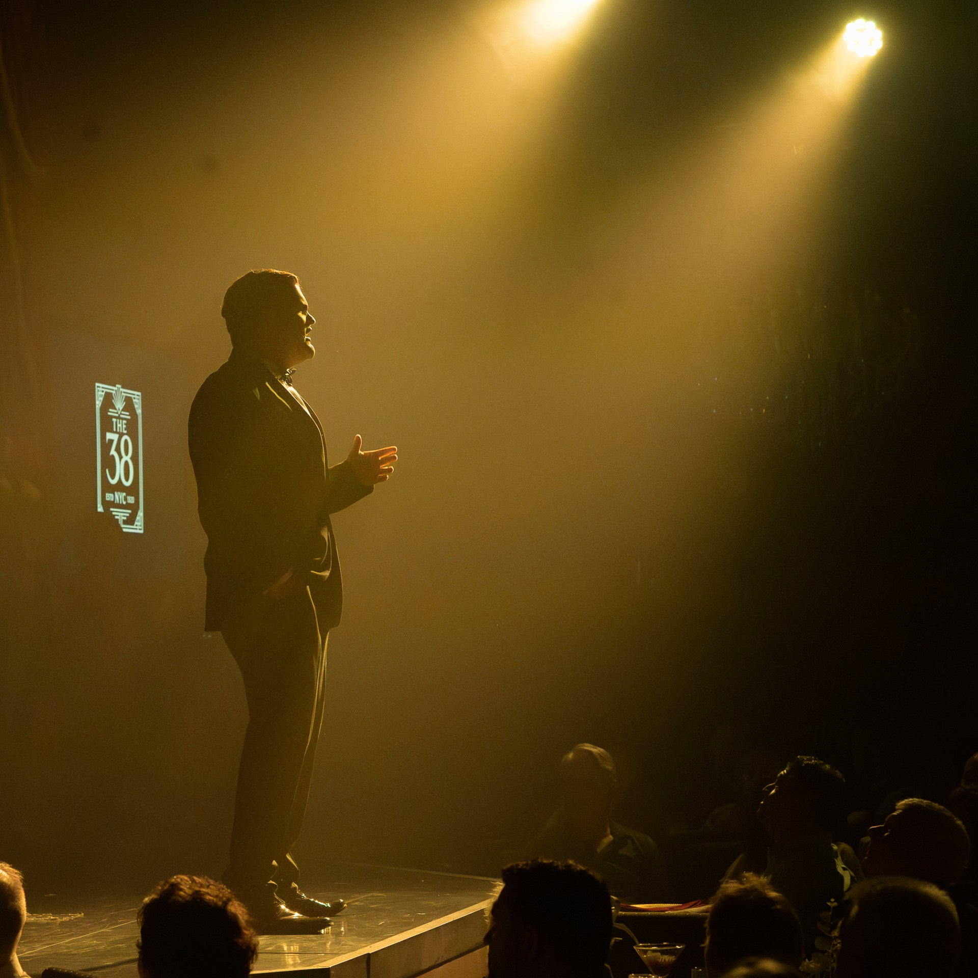 Silhouette of a man speaking on stage under bright spotlight to an audience.