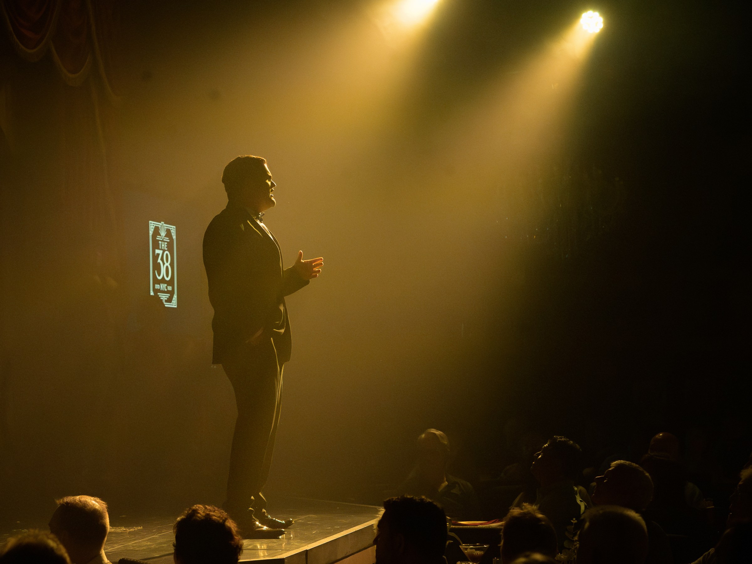 Silhouette of a man speaking on stage under bright spotlight to an audience.