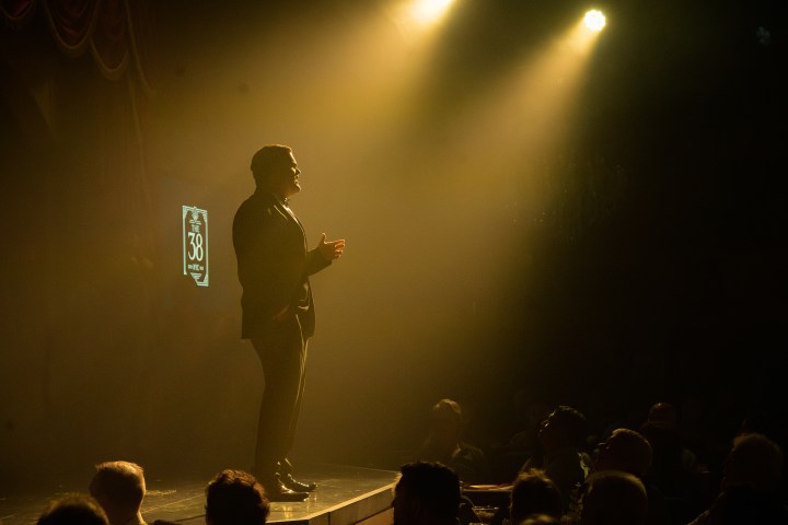 Silhouette of a man speaking on stage under bright spotlight to an audience.