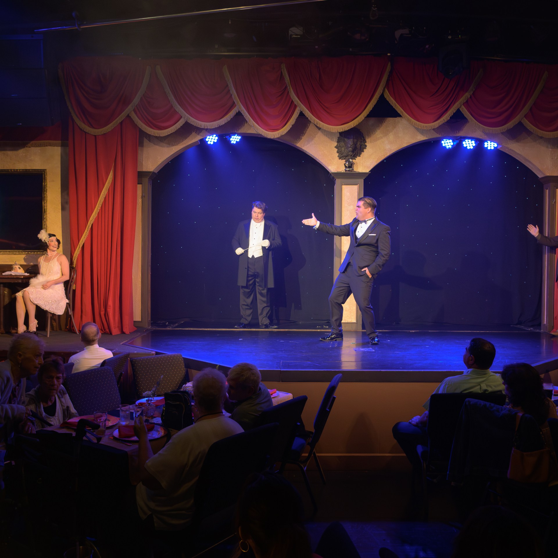 Theater scene with performers in period costumes on stage, audience seated at tables.