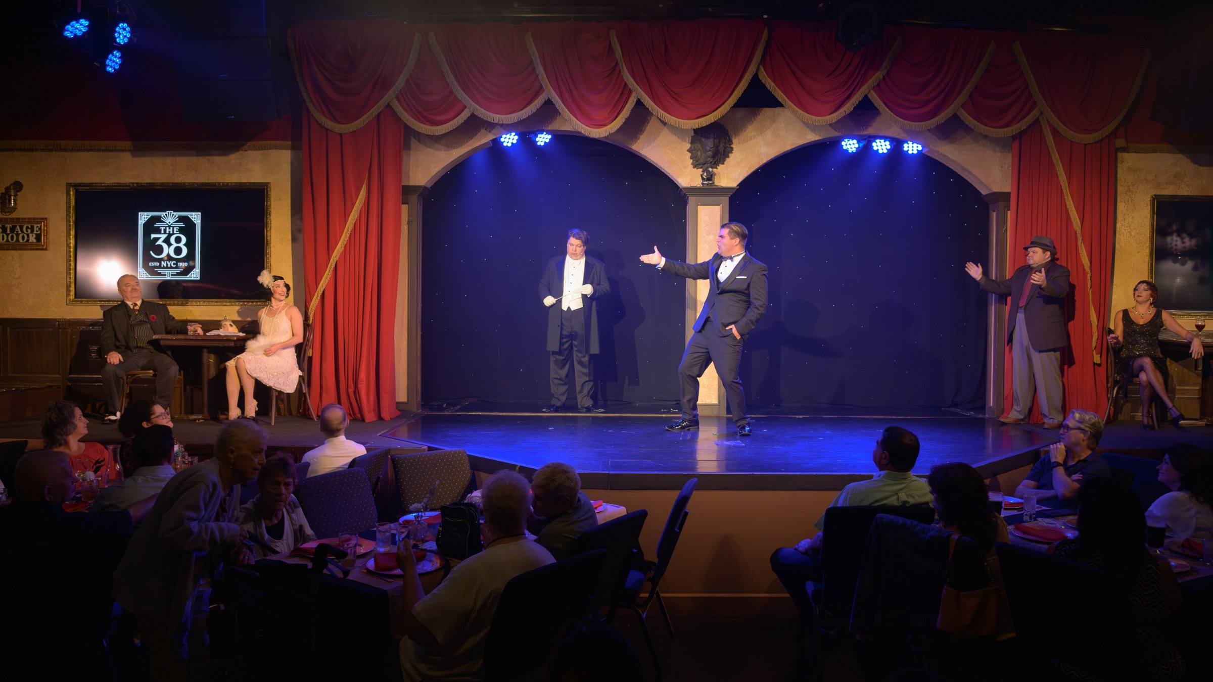 Theater scene with performers in period costumes on stage, audience seated at tables.