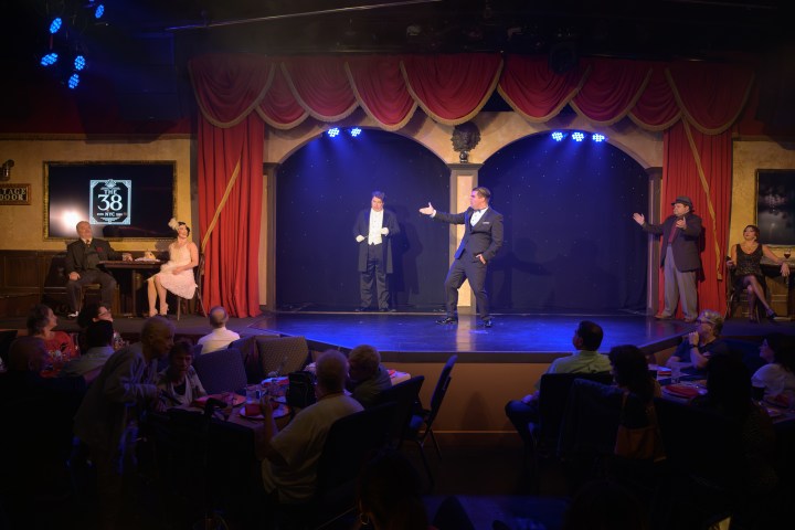 Theater scene with performers in period costumes on stage, audience seated at tables.