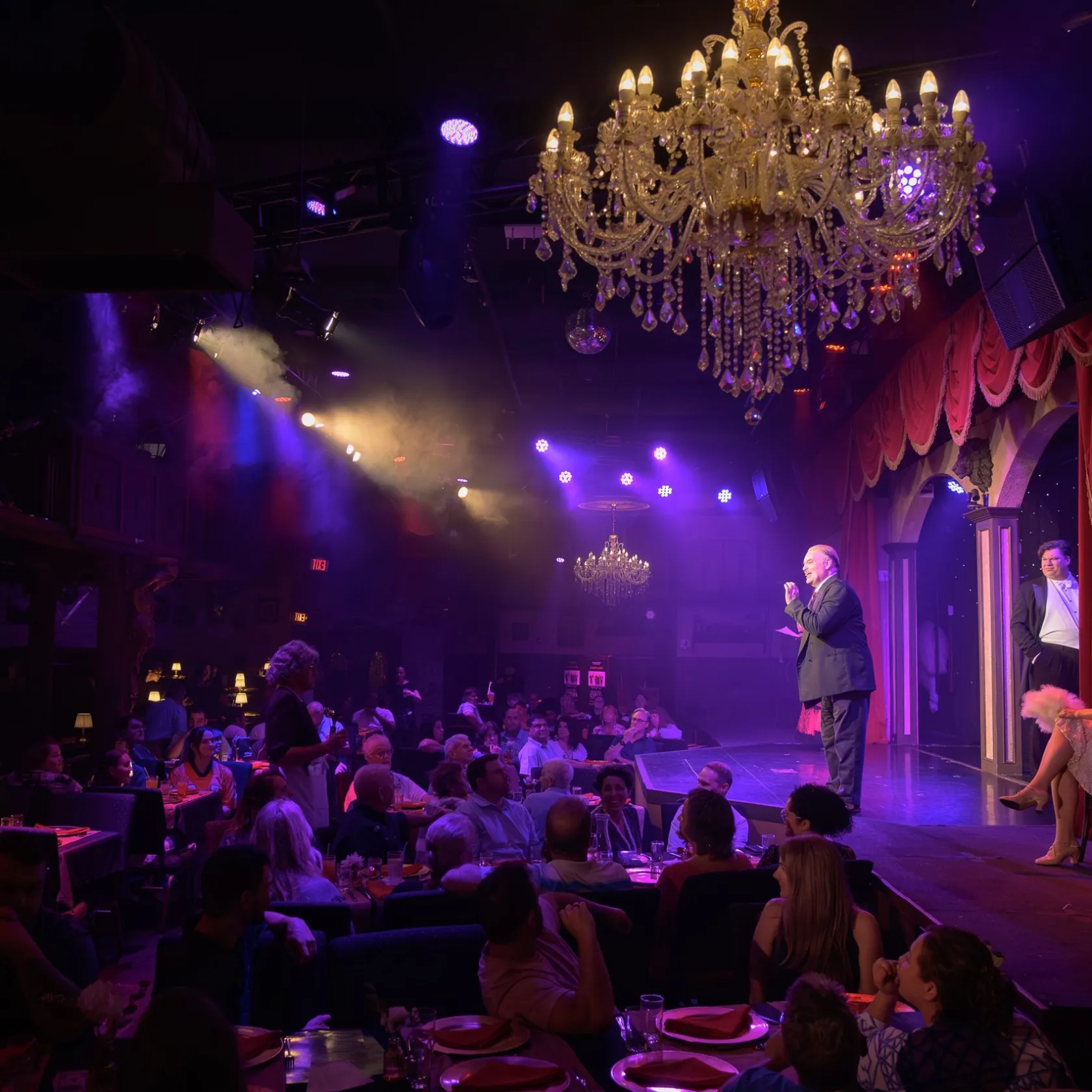 Cabaret show with performers on stage, audience seated, and chandeliers overhead in a dimly lit venue.
