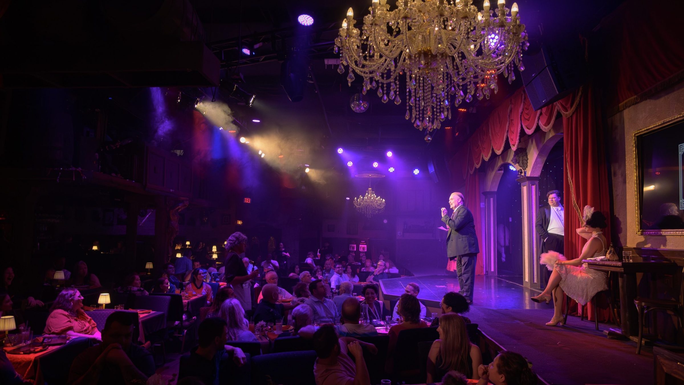 Cabaret show with performers on stage, audience seated, and chandeliers overhead in a dimly lit venue.