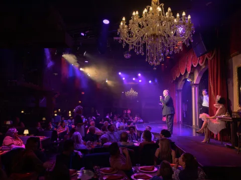 Cabaret show with performers on stage, audience seated, and chandeliers overhead in a dimly lit venue.