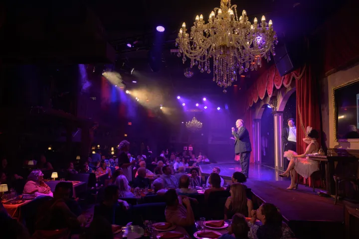 Cabaret show with performers on stage, audience seated, and chandeliers overhead in a dimly lit venue.