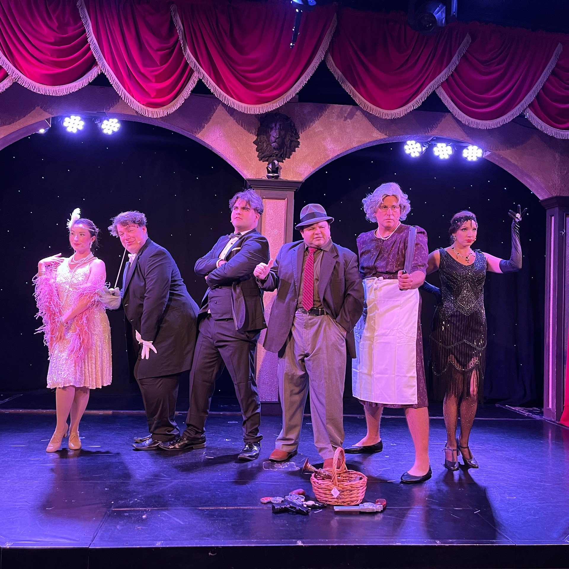 Six actors in vintage costumes pose dramatically on a stage with red curtains.