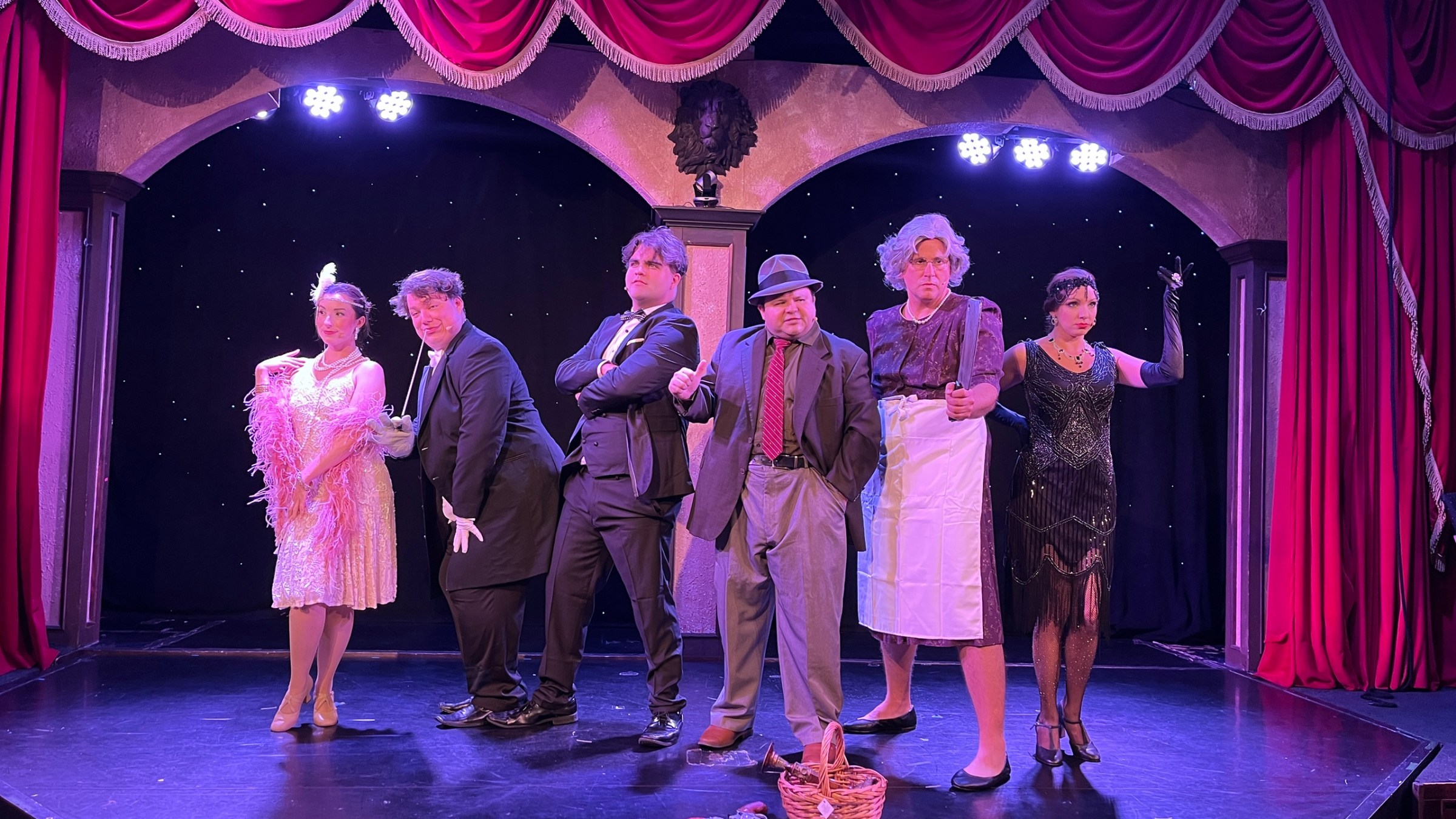 Six actors in vintage costumes pose dramatically on a stage with red curtains.