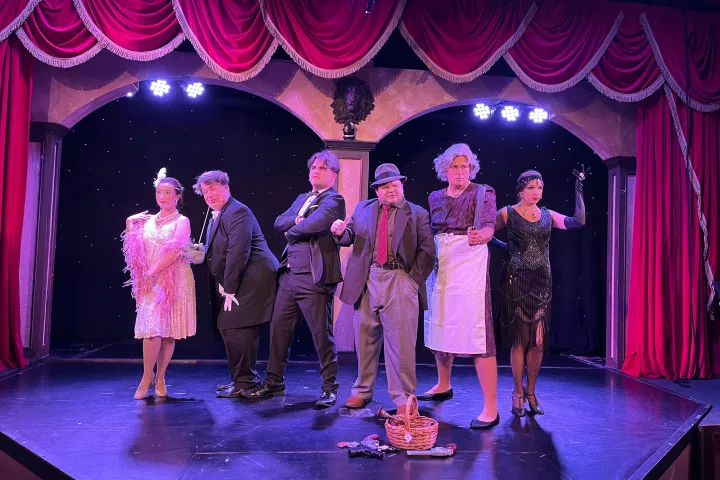 Six actors in vintage costumes pose dramatically on a stage with red curtains.