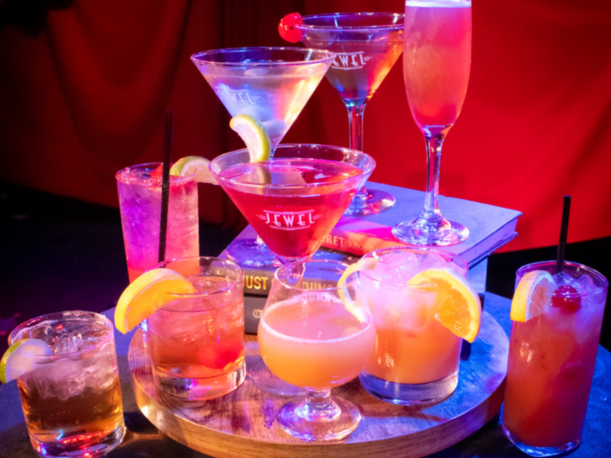Assorted cocktails on a tray with orange slices and cherries, set against a red background.