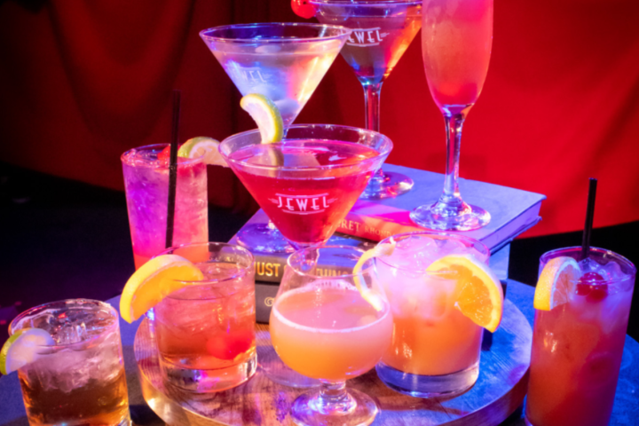 Assorted cocktails on a tray with orange slices and cherries, set against a red background.