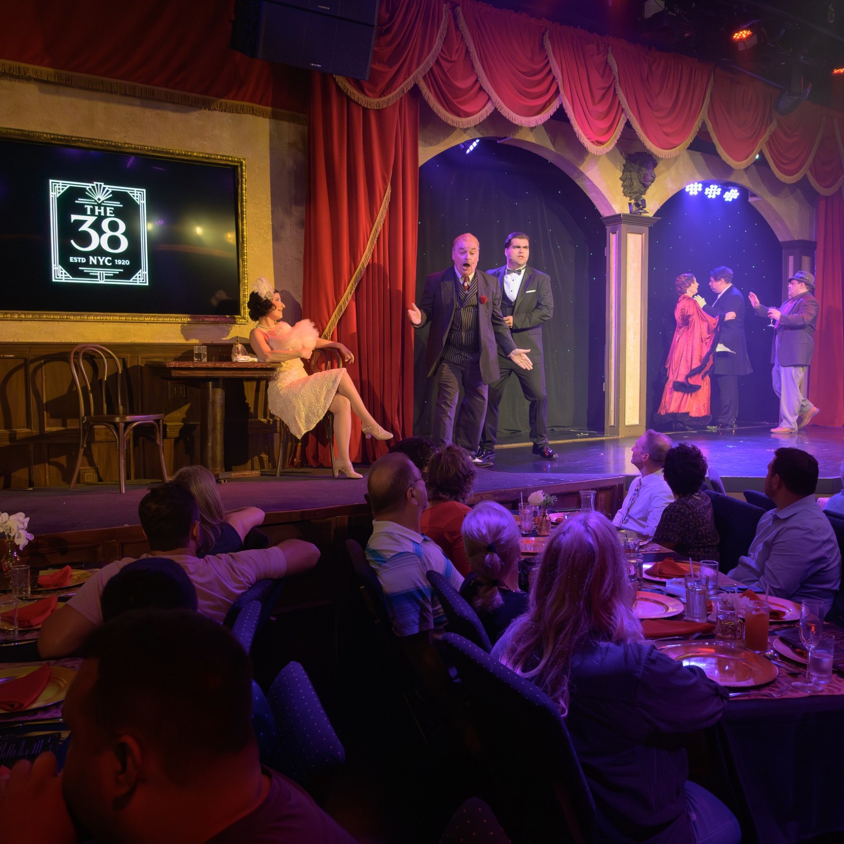 Actors performing on stage as audience watches in a dinner theater setting.