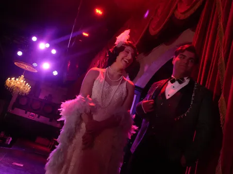 Two people in vintage attire on stage with red lighting and a chandelier.