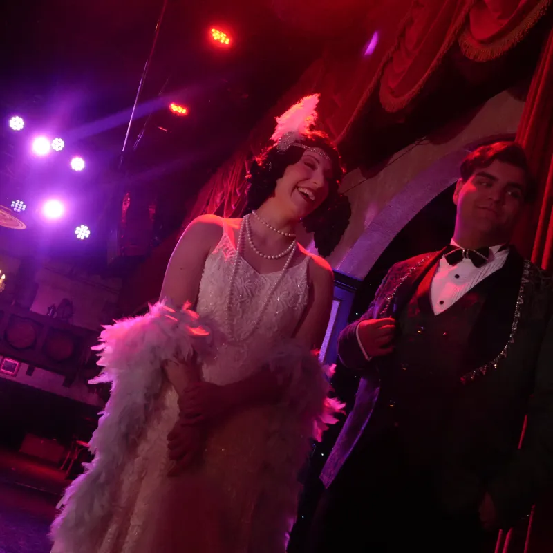 Two people in vintage attire on stage with red lighting and a chandelier.