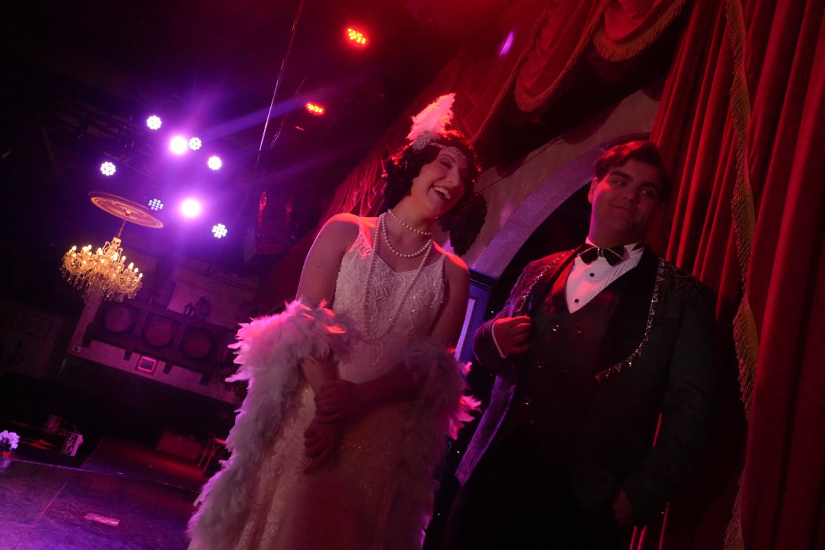 Two people in vintage attire on stage with red lighting and a chandelier.