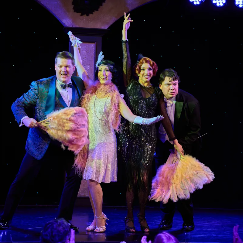 Four performers in 1920s-themed attire pose on stage with feather fans.