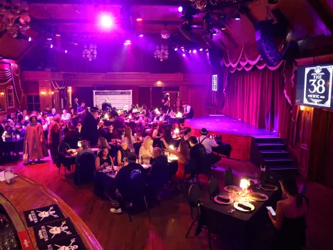 People seated at tables in a dimly lit event venue with a stage and purple lighting.
