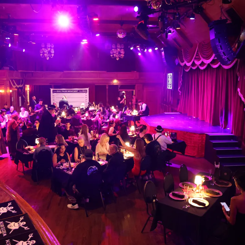 People seated at tables in a dimly lit event venue with a stage and purple lighting.