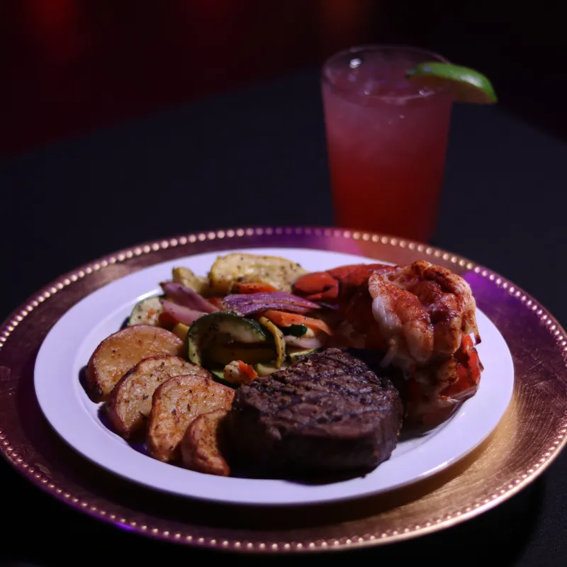 Plate with steak, shrimp, vegetables, and a drink with lime on the side.