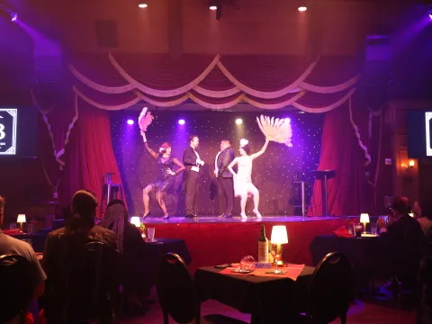 Cabaret performers on stage with feather fans and audience seated at tables.