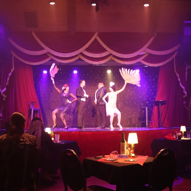 Cabaret performers on stage with feather fans and audience seated at tables.